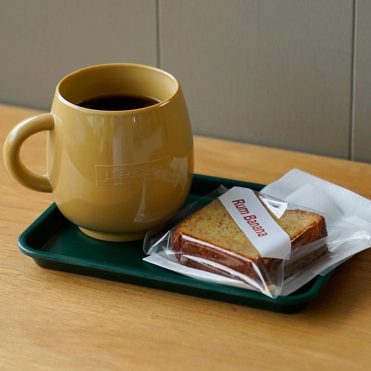 Een Penco Mok Beige gevuld met zwarte koffie staat op een groen dienblad naast verpakt bananenbrood met rum en een gevouwen wit servet, allemaal op een houten tafel die doet denken aan Japans design met een retro-look.