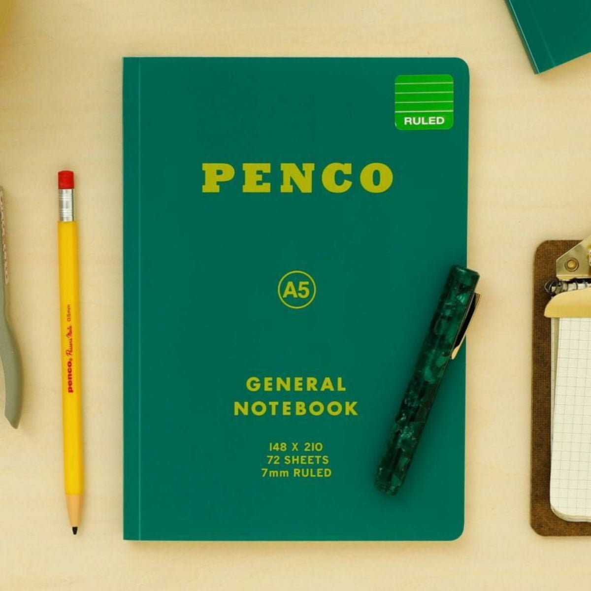 Een Penco Notitieboek Groen A5 met een pen, een geel potlood met rode gum en een klein klembord met papier liggen netjes gerangschikt op een licht houten oppervlak.