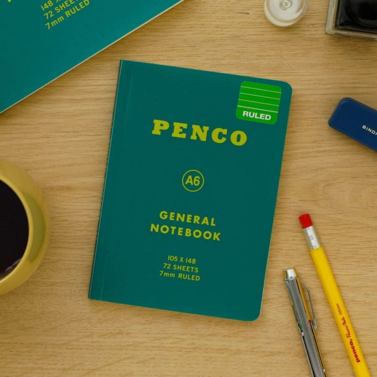Een Penco Notitieboek Groen A6 rust op een houten bureau met een koffiekopje, geel potlood, rode pen, mechanisch potlood en groenblauw notitieboek. Het Penco notitieboek heeft een groene Ruled sticker op de kaft.