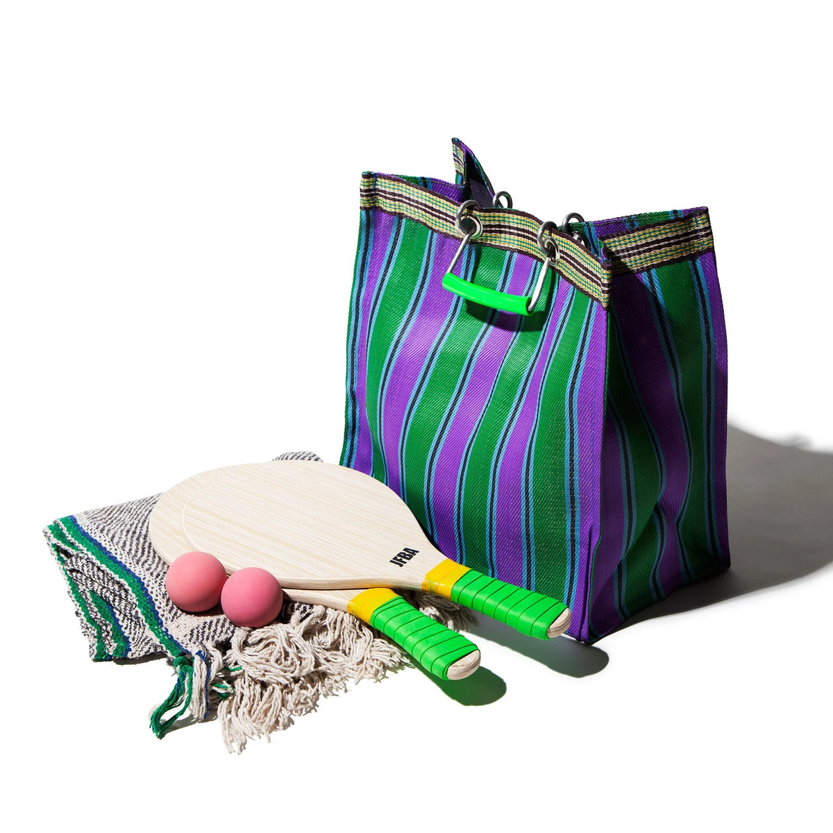 De Puebco Gerecycleerde Tas Strepen Groen Paars Vierkant, een kleurrijke gerecyclede tas van Puebco, rust op een geweven deken met twee houten paddle rackets (groene handvatten) en twee roze ballen - alles tegen een witte achtergrond.