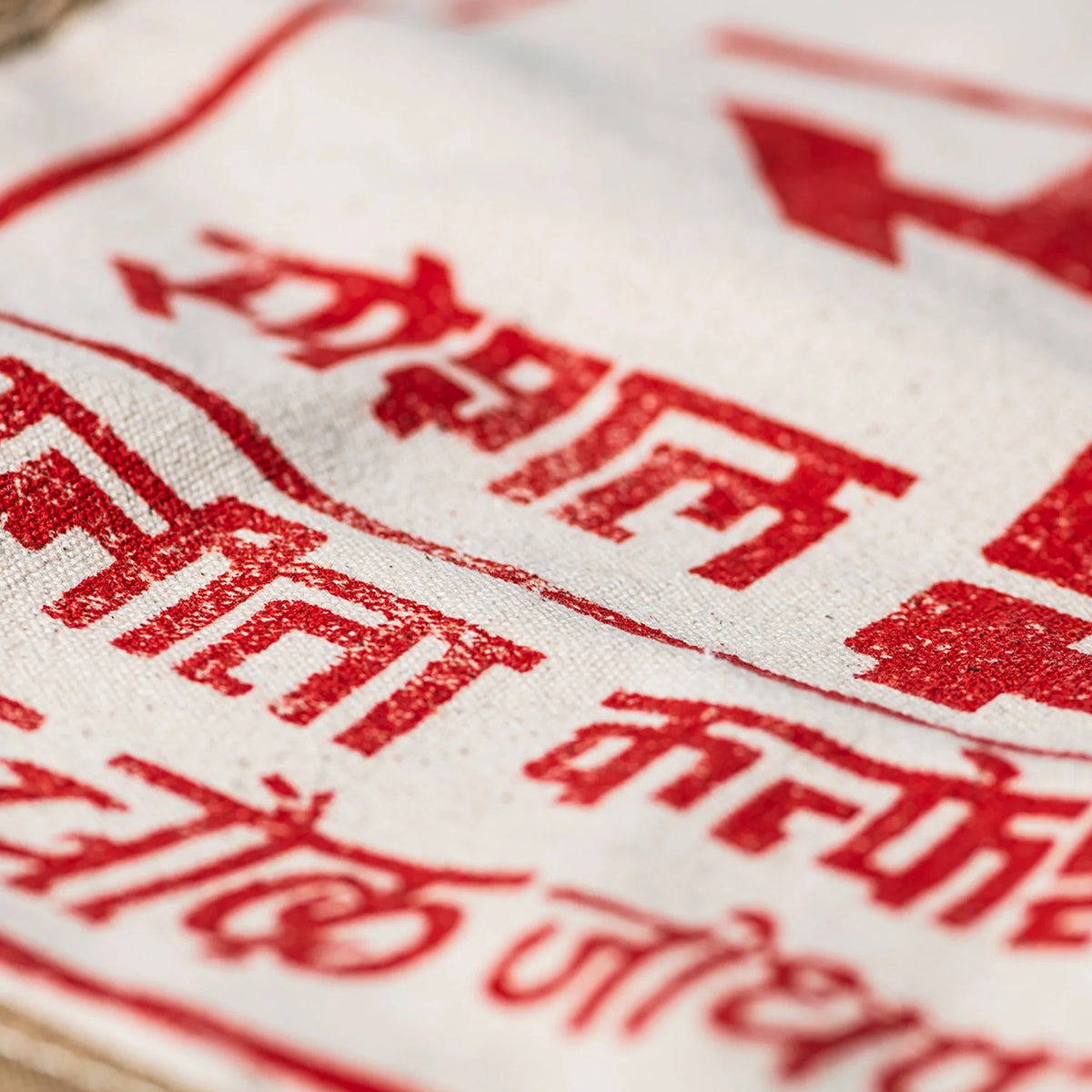 Close-up van rode Hindi tekst op een witte, licht gerimpelde Puebco India Souvenir Tas. De textuur van de katoenen stof en de inktdetails van Puebco zijn duidelijk zichtbaar, wat deze tas uniek maakt.