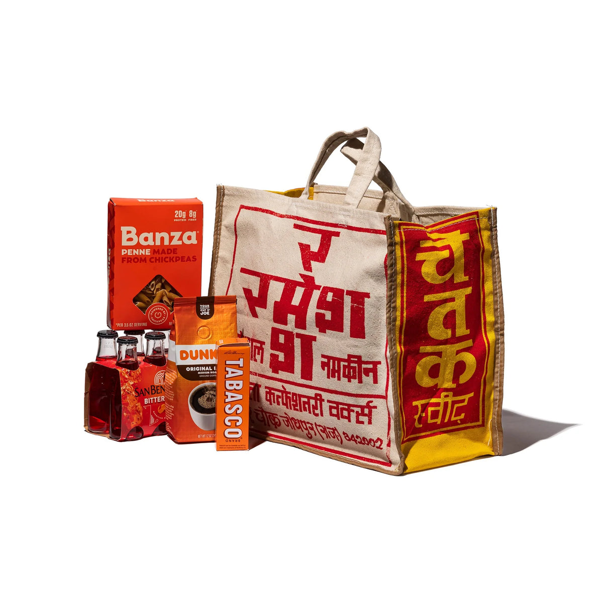 De Puebco India Souvenir Tas heeft vetgedrukte rode en gele Hindi tekst, omringd door Banza penne, Dunkin koffie, San Benedetto drankjes, Tabasco saus en Walkers zandkoekjes op een witte achtergrond - perfect om cadeau te geven.