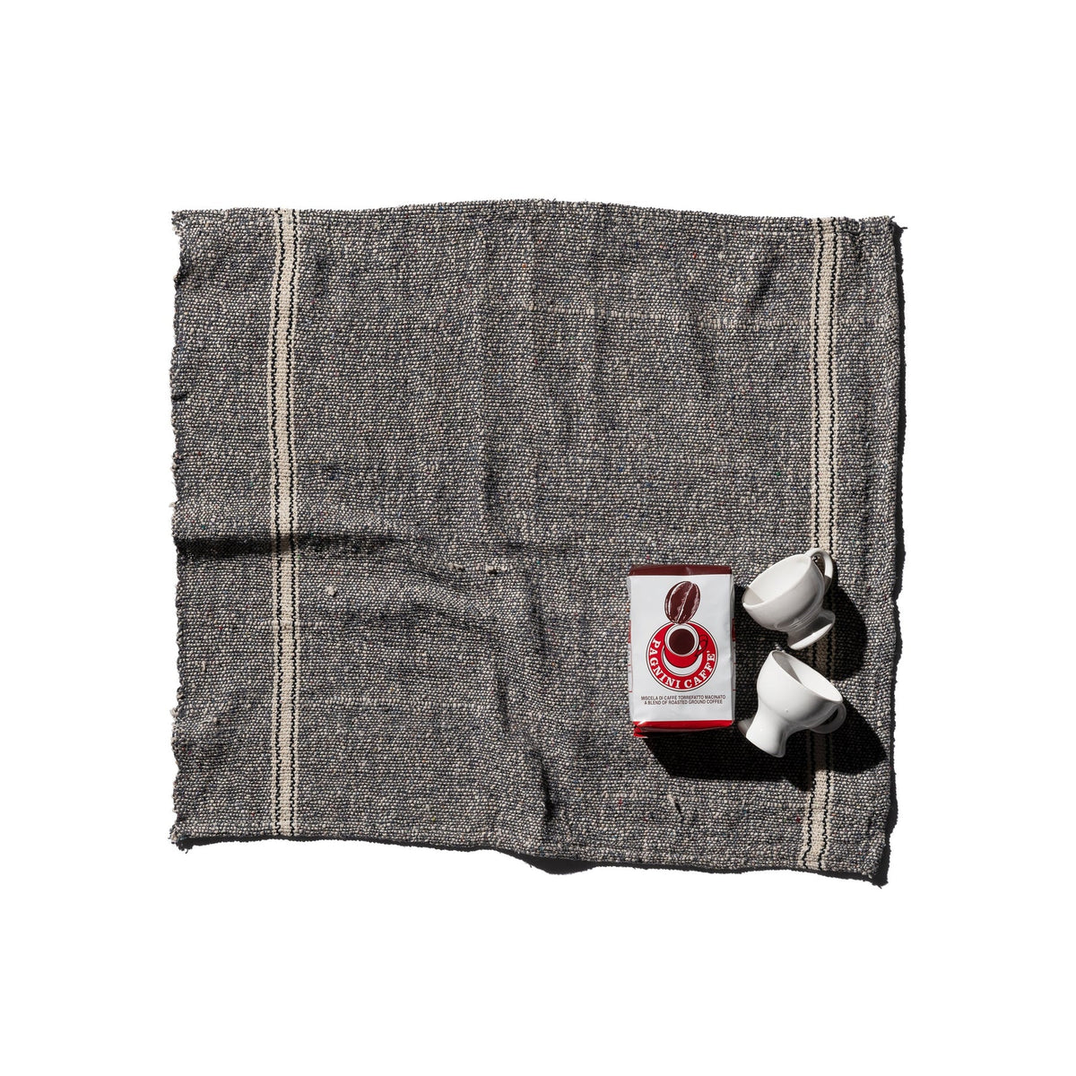 De Puebco India Theedoek Grijs 60/60 van Puebco, vervaardigd uit gerecycleerde stoffen, vormt het decor voor twee witte koffiekopjes en een rood-witte Counter Culture Coffee verpakking tegen een witte achtergrond.