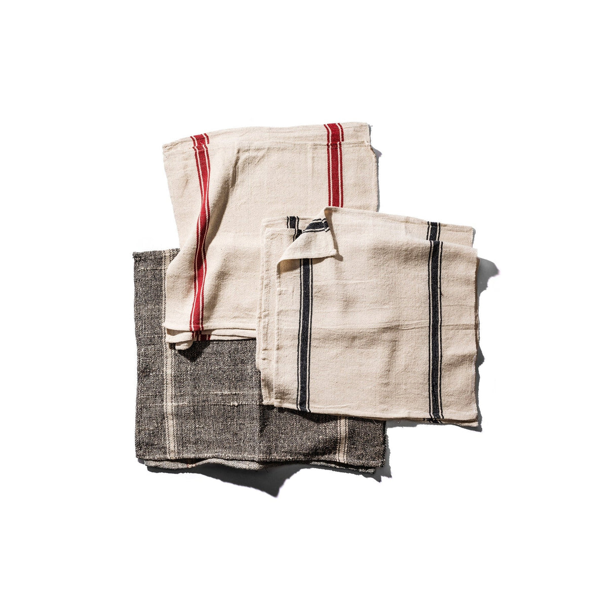 Drie gevouwen Puebco India Theedoek Zwart 60/60 servetten - beige met rode strepen, beige met zwarte strepen en donkergrijs (Theedoek Zwart) met witte strepen - zijn gemaakt van gerecycleerde stoffen in India en liggen op een witte achtergrond.