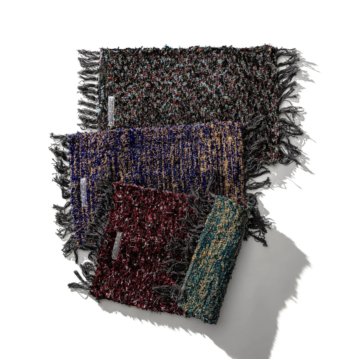 Drie veelkleurige geweven sjaals met textuur en franjes zijn diagonaal gerangschikt op een witte achtergrond, elk met een uniek patroon. Hun ambachtelijke stijl doet denken aan de Puebco Mat Gerecycleerd Textiel 63/41 van Puebco. Schaduwen naar rechts.