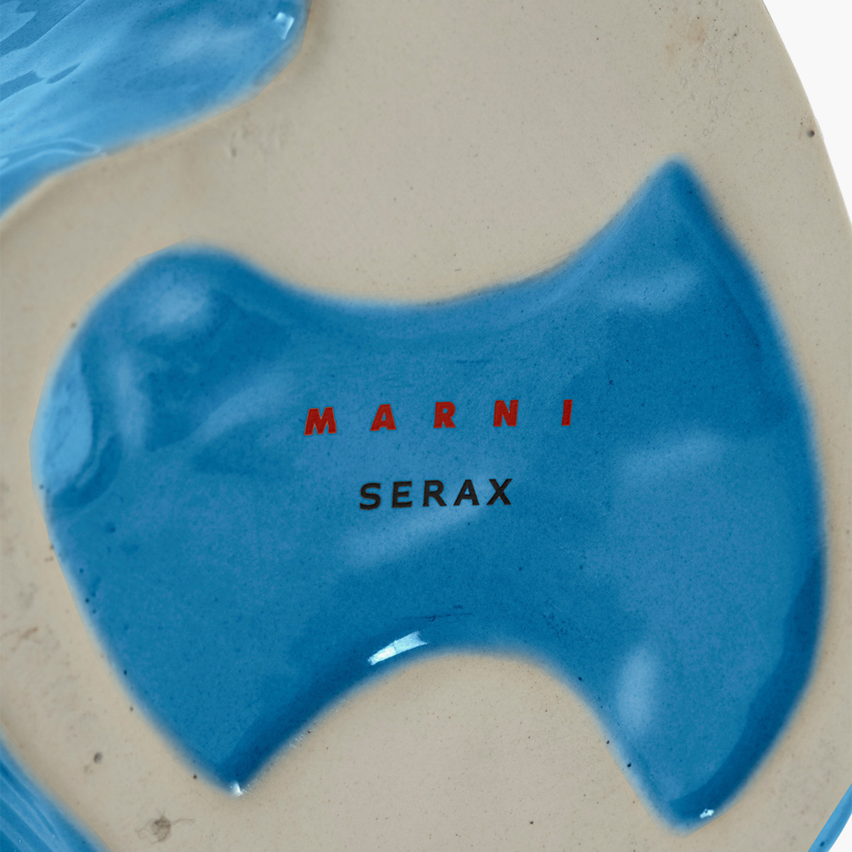 Close-up van de Serax Marni After Midnight Uil Vaas Blauw S met een glanzend blauw abstract ontwerp en "MARNI" in rood, "SERAX" in zwart in het midden. De blauwe vaas van keramiek staat tegen een gebroken witte gestructureerde achtergrond.