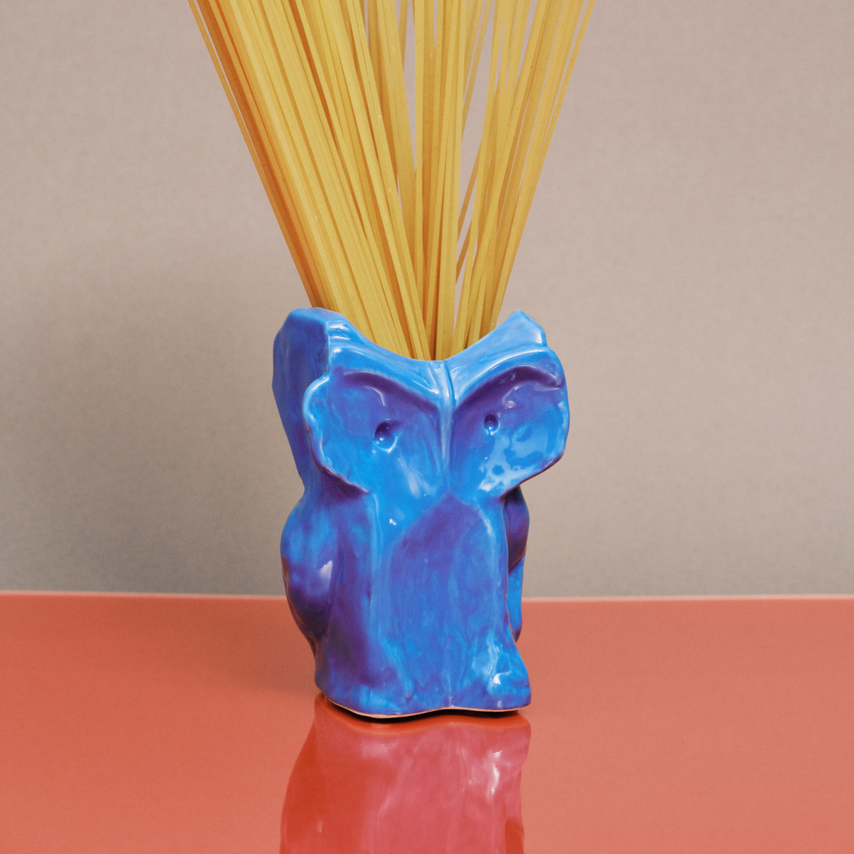 De Serax Marni After Midnight Uil Vaas Blauw XS, een vaas van blauw keramiek van Serax, houdt ongekookte spaghetti op een glanzend koraaloppervlak tegen een beige achtergrond.
