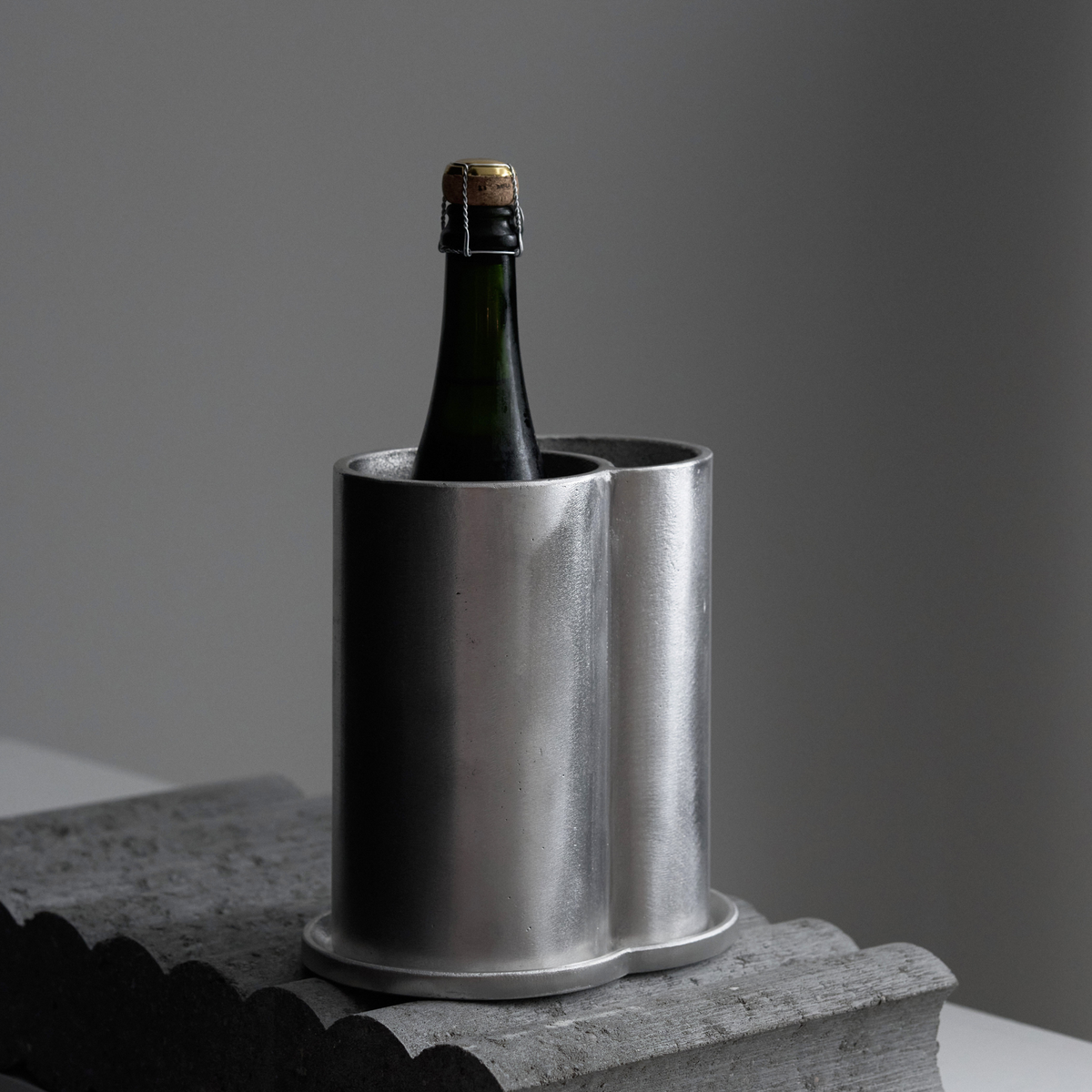 In de strakke, zilverkleurige Serax Surface Wijnkoeler van Serax, elegant vervaardigd uit verfijnd aluminium, staat een groene wijnfles met een kurk. De wijnkoeler staat op een grijs oppervlak met structuur, tegen een effen, zacht verlichte achtergrond.