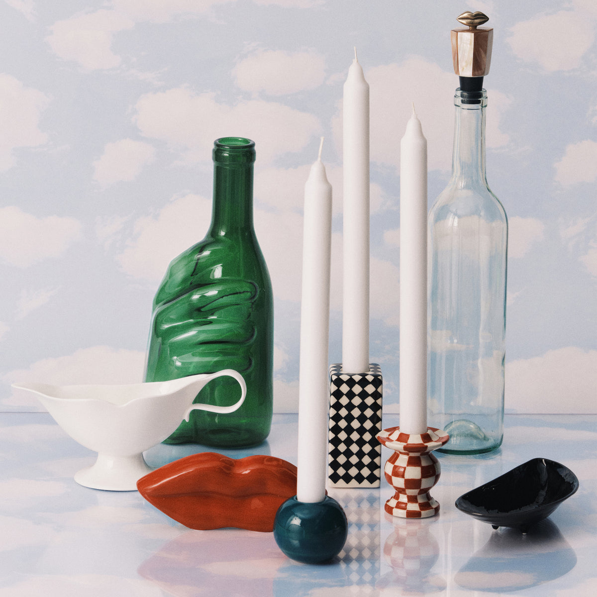 Een stilleven toont decoratieve objecten, waaronder twee flessen, een witte juskom en de Serax Wouters & Hendrix Edward Karaf Groen set tussen kaarsen en schalen tegen een achtergrond met een hemelthema.