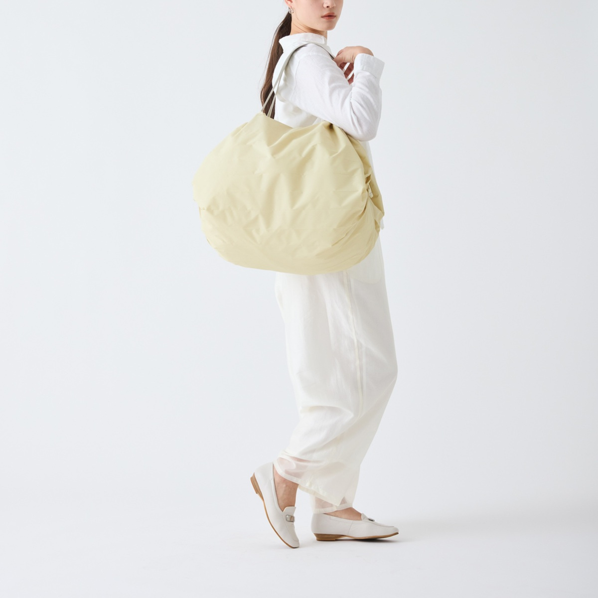 Een vrouw in witte kleding en loafers draagt een grote, beige Shupatto Opvouwbare Boodschappentas Cashew Nuts L over haar schouder, terwijl ze tegen een effen witte achtergrond staat.