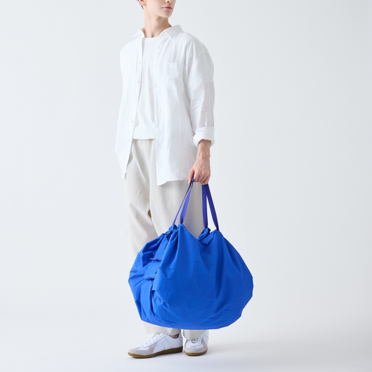 Een persoon in een los wit overhemd, een lichte broek en witte sneakers houdt een Shupatto Opvouwbare Boodschappentas Clear Sky Blauw L-draagtas met twee hengsels vast tegen een effen witte achtergrond.