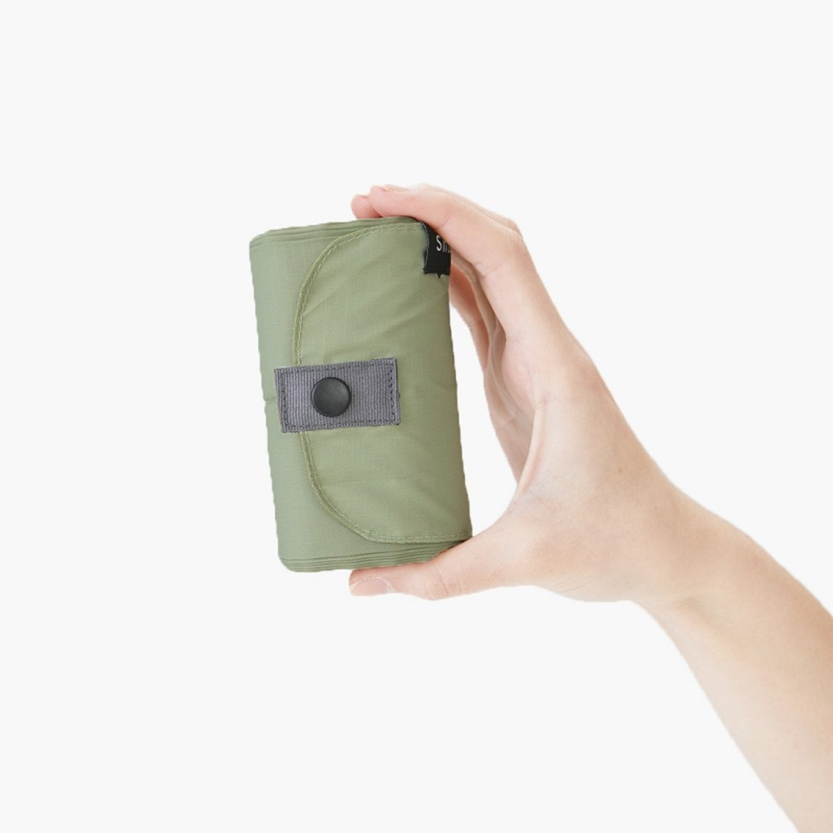 Op een effen witte achtergrond is een hand te zien die een Shupatto Opvouwbare Boodschappentas Forest Groen L vasthoudt, compact opgerold in het olijfgroene etui met een grijze klep met knoop, van het merk Shupatto.