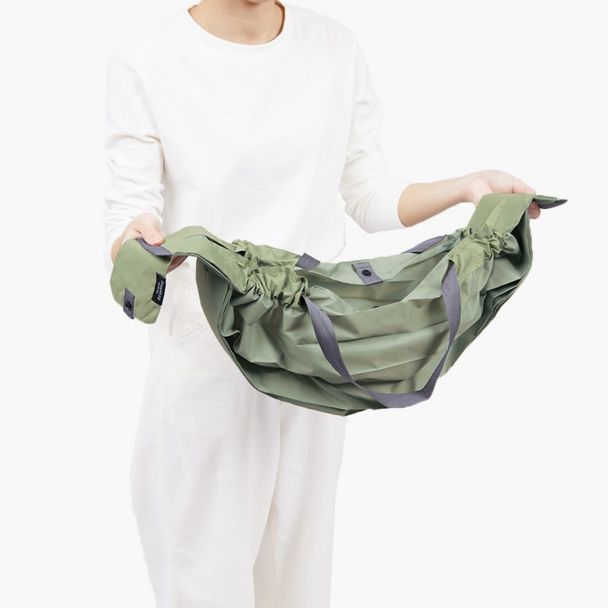 Een persoon in een witte outfit houdt de Shupatto Opvouwbare Boodschappentas Brick Bruin L vast, waardoor het flexibele en ruime ontwerp goed tot zijn recht komt tegen een effen witte achtergrond.