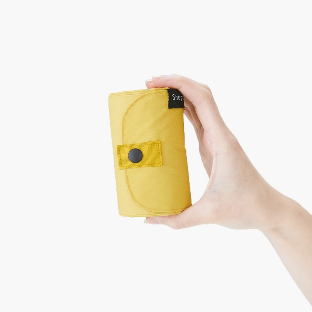 Op een effen witte achtergrond is een hand te zien die een compacte, opgerolde Shupatto Opvouwbare Boodschappentas Mosterd L vasthoudt, voorzien van een drukknoopsluiting en een zwart label.
