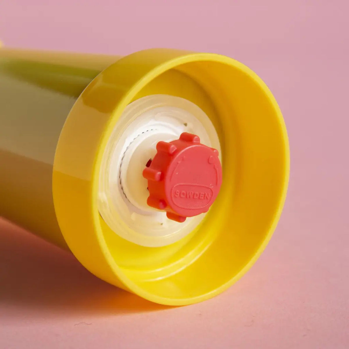 Een close-up van de gele dop en rode draaizegel van de Sowden Zout-en Pepermolen Geel van Sowden, tegen een roze achtergrond. Op de speelse rode zegel, geïnspireerd op de ontwerpen van George Sowden, staat "ZWEDEN" in reliëf.