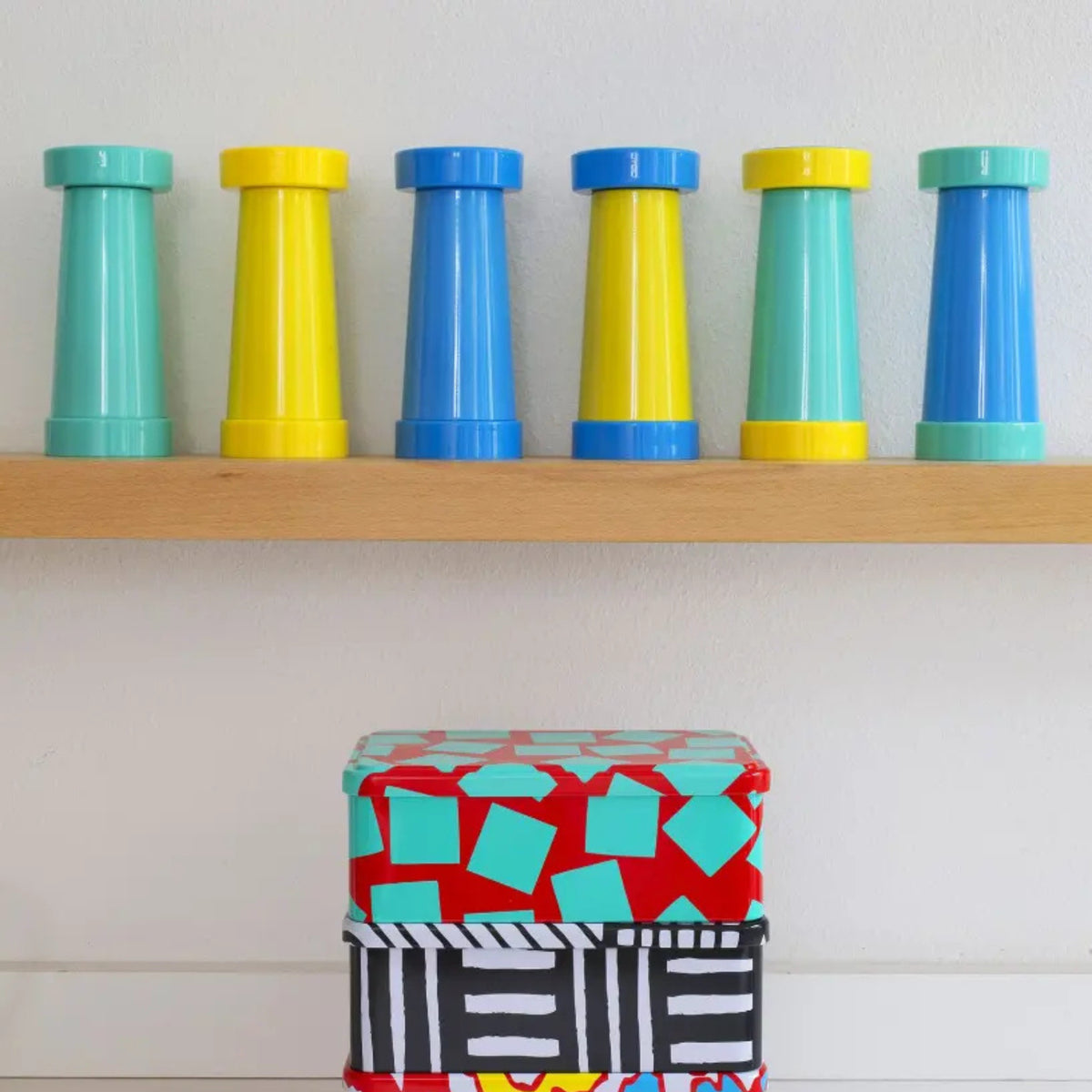 Zes gele Zout-en Pepermolen van Sowden, elk in de vorm van een schaaktoren, staan op een houten plank. Eronder staan twee decoratieve dozen met gedurfde patronen - een in rood, turquoise en wit - voor een speels contrast.