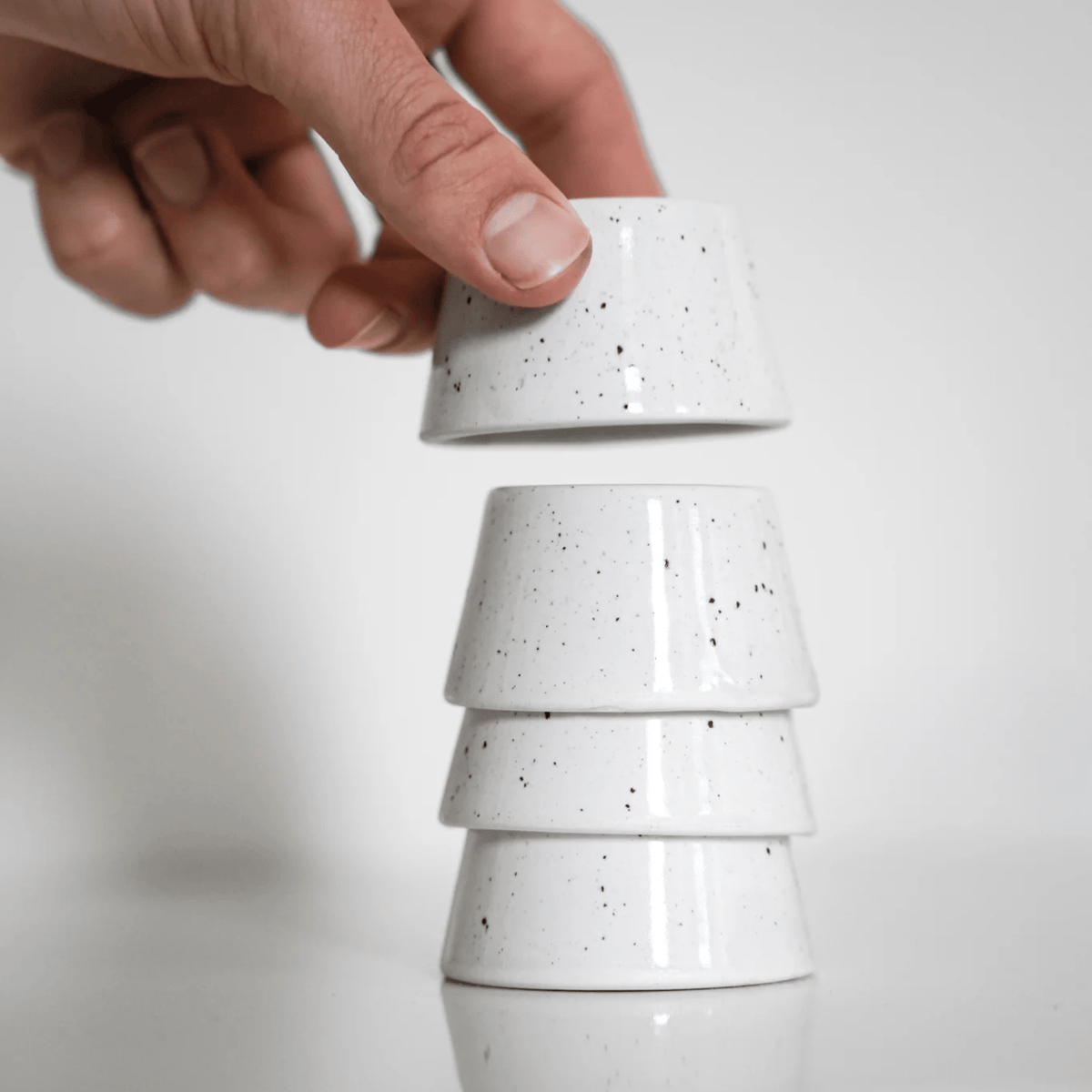 Een hand tilt een Stock Antwerpen Eierdopje Wit Spikkel - een klein, wit, gespikkeld eierdopje van Stock Antwerpen - van een stapel van drie tegen een effen witte achtergrond.