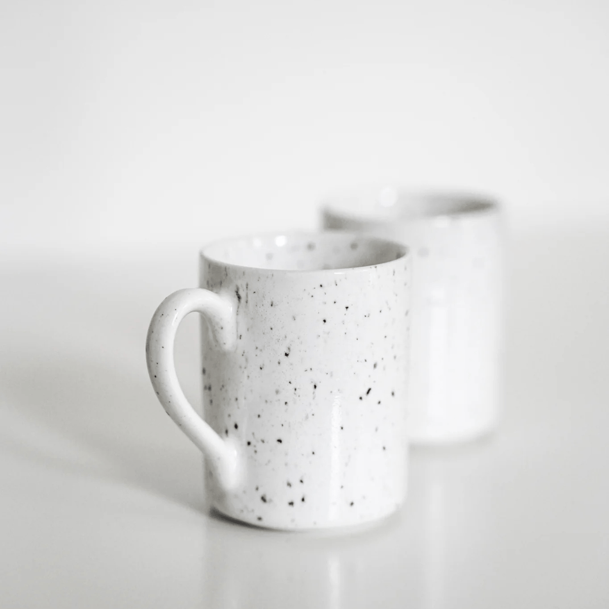Twee Stock Antwerpen Espresso kop Wit Spikkel mokken van Stock Antwerpen zijn geplaatst op een wit vlak. Het handvat van de mok op de voorgrond is duidelijk zichtbaar, terwijl de andere mok een beetje wazig is achteraan in dit minimalistische tafereel.