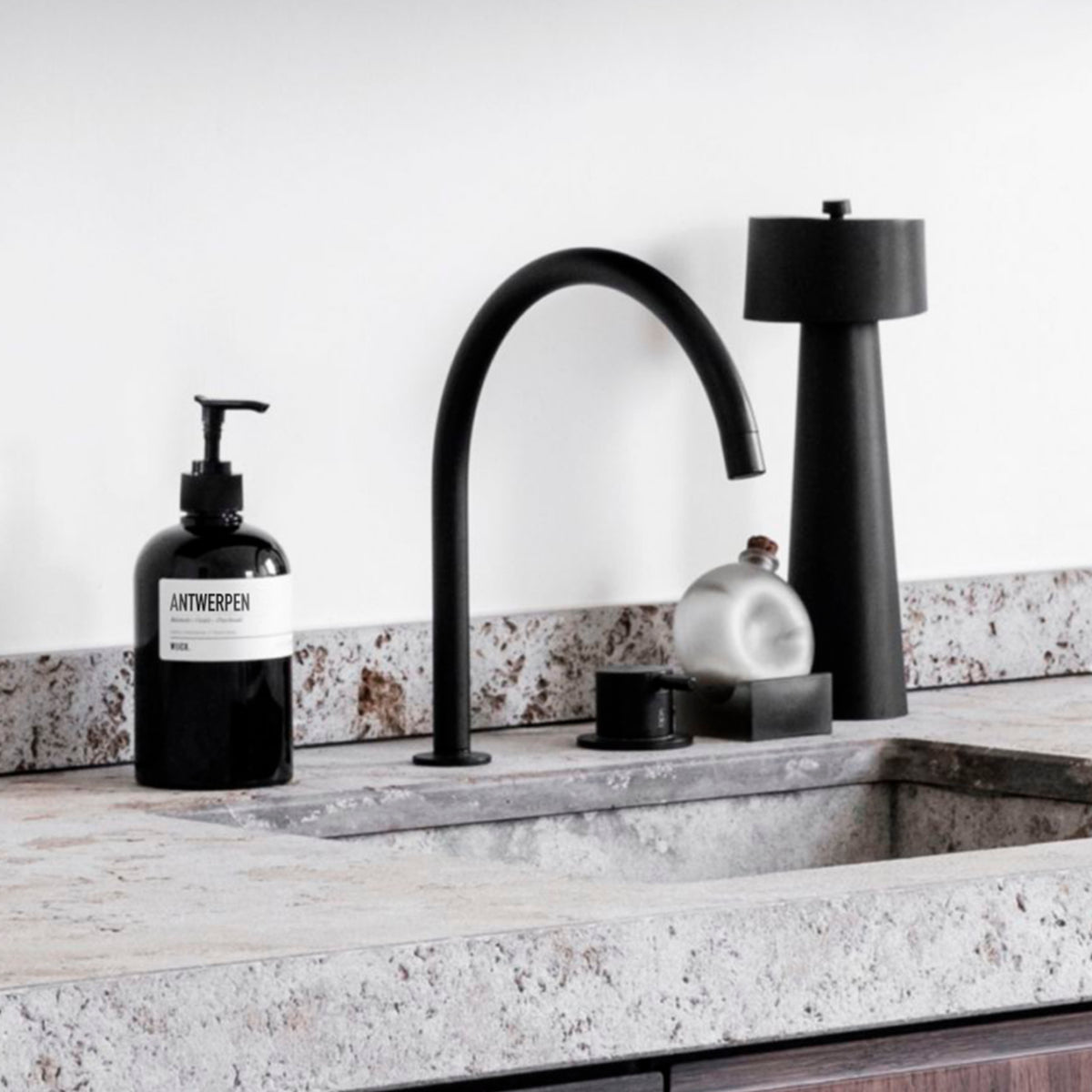Een modern aanrecht met een minimalistische zwarte kraan, een Wijck Handzeep Boulevard Antwerpen zeepfles van 500 ml, een matzwarte lamp en decor - alles op een licht stenen oppervlak en achterwand.
