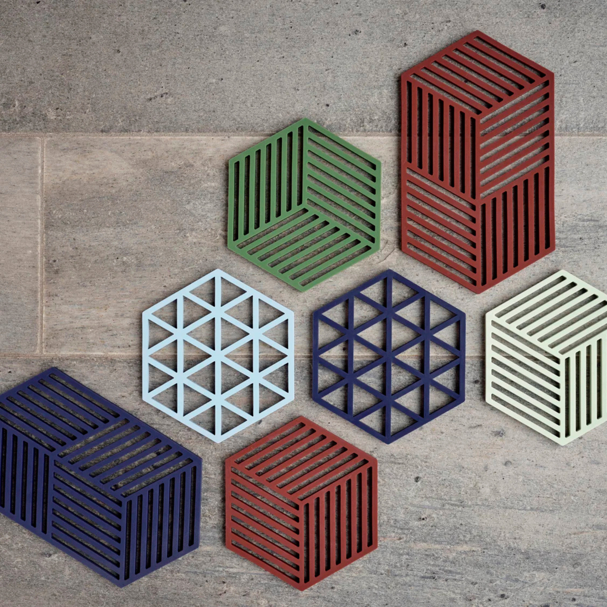 De Zone Denmark Hexagon Trivet XL Midnight van Zone Denmark heeft opvallende geometrische patronen in levendige kleuren op een grijze ondergrond – vier met strepen, drie met driehoekige rasters – en is ideaal als onderzetter of stijlvolle siliconen onderzetter.