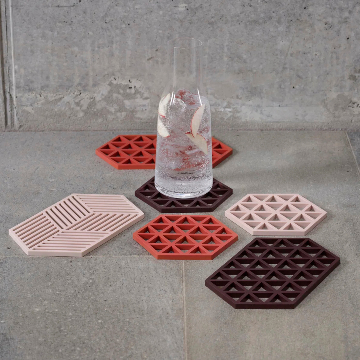 Op een grijze tegelvloer staat een groot glas bruisend water met ijs en appelschijfjes, omringd door zes Zone Denmark Hexagon Trivet XL Roze onderzetters in tinten roze, rood en bordeauxrood.