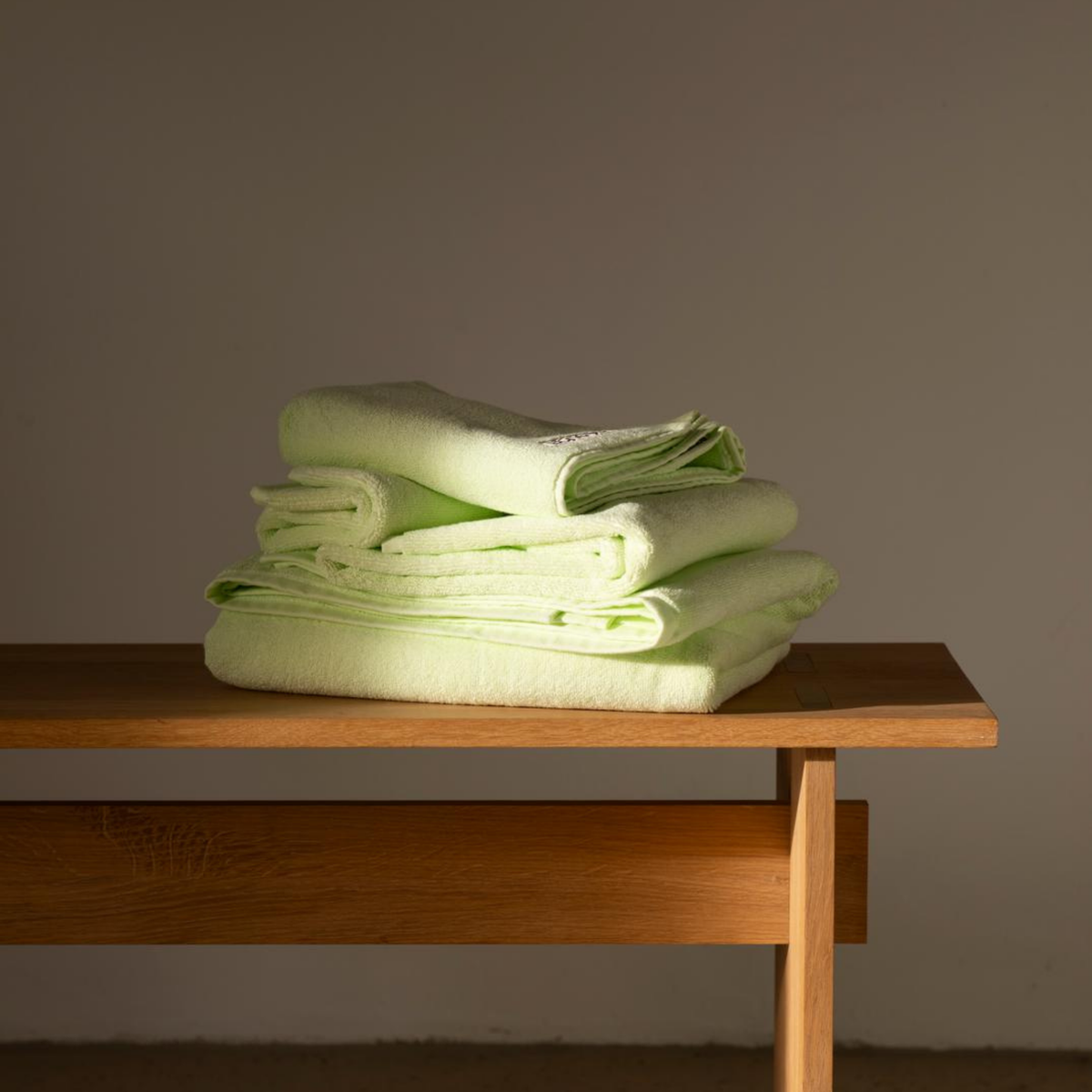 Een netjes opgevouwen stapel Bongusta Naram Badhanddoek Lemon Sorbet 70/140 ligt op een eenvoudige houten bank tegen een effen, lichtgekleurde muur.