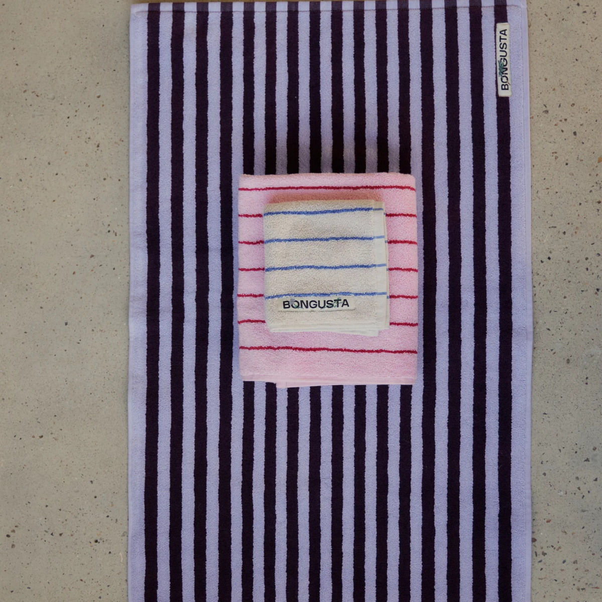 Een grote gestreepte Bongusta Naram Badmat Wine & Lavender Blue 50/80 van biologisch katoen ligt op een gespikkeld oppervlak met een kleinere roze handdoek erop gevouwen. Beide zijn voorzien van BONGUSTA labels - perfect voor de badkamer.