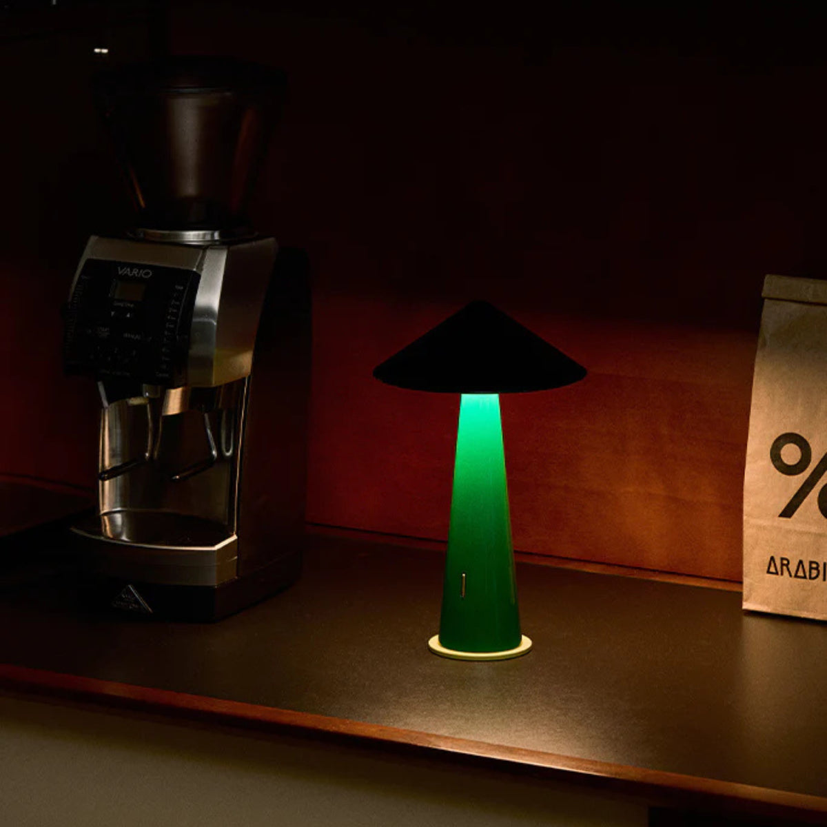 Een Fine Lumens Tipsy Lamp LED Oplaadbaar Zwart Emerald Groen werpt een gloed op een donker aanrecht naast een koffiemolen en een bruine papieren zak met het label "% ARABICA".