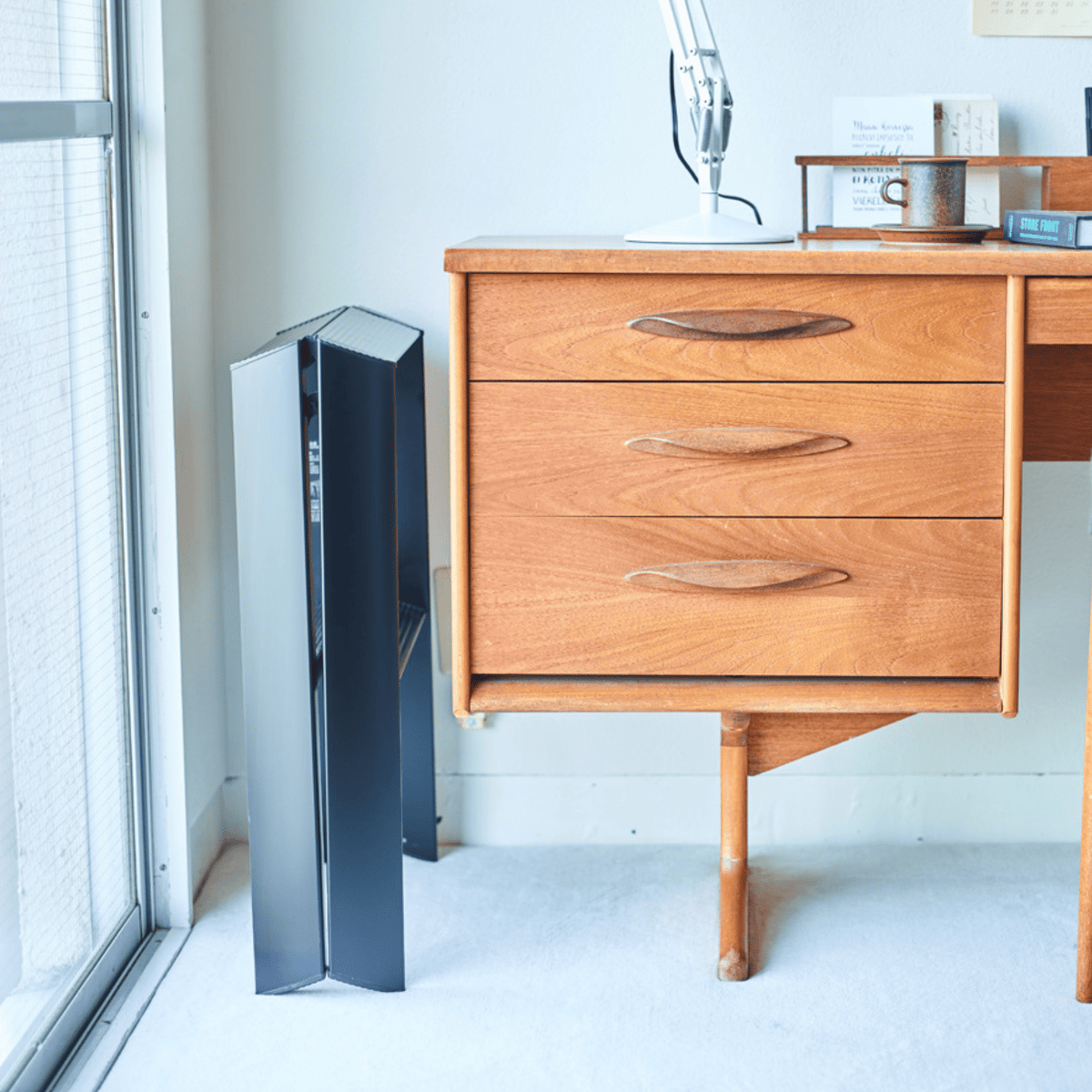 De Hasegawa Lucano Ladder 2 Stap Zwart staat strak en rechtop naast een mid-century houten bureau met drie laden, een witte lamp, beker en papieren. Zacht natuurlijk licht benadrukt het minimalistische Japanse ontwerp van Hasegawa.