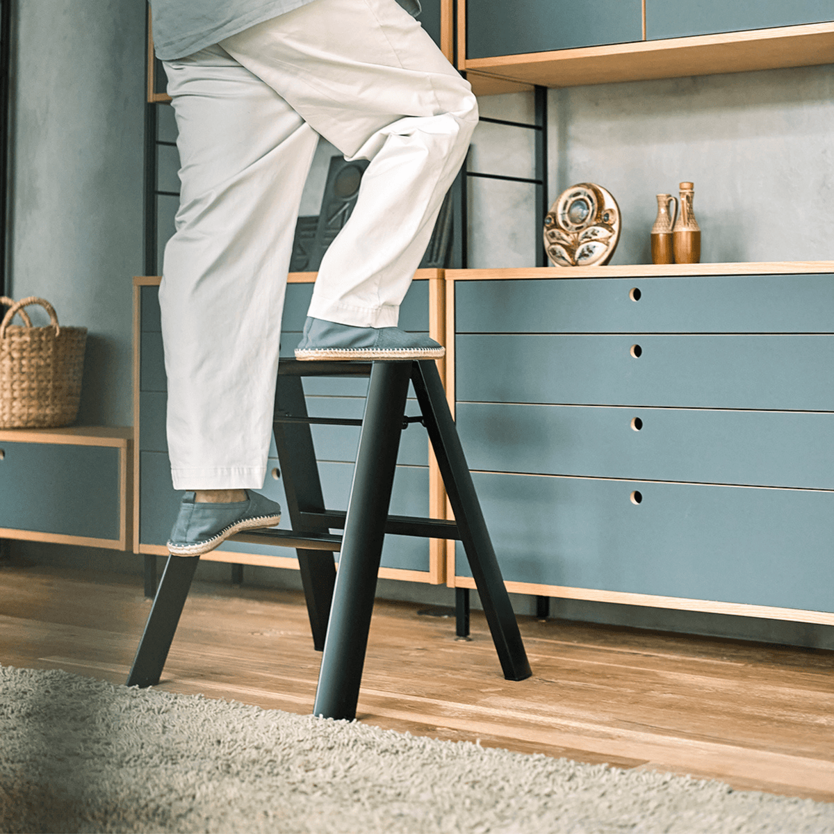 Een persoon in een witte broek en slippers staat op een Hasegawa Lucano Ladder 2 Step Zwart van Hasegawa in een moderne, op Japan geïnspireerde woonkamer met blauwe kasten, houten planken en decoratieve voorwerpen op de achtergrond.