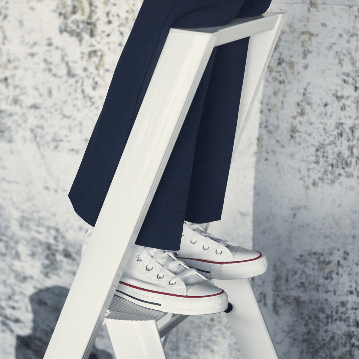 Een persoon in een donkere broek en witte sneakers met rode en blauwe accenten staat op een Hasegawa Lucano Ladder 3 Step Wit van Hasegawa, ontworpen in Japan, tegen een lichtgekleurde muur met textuur.