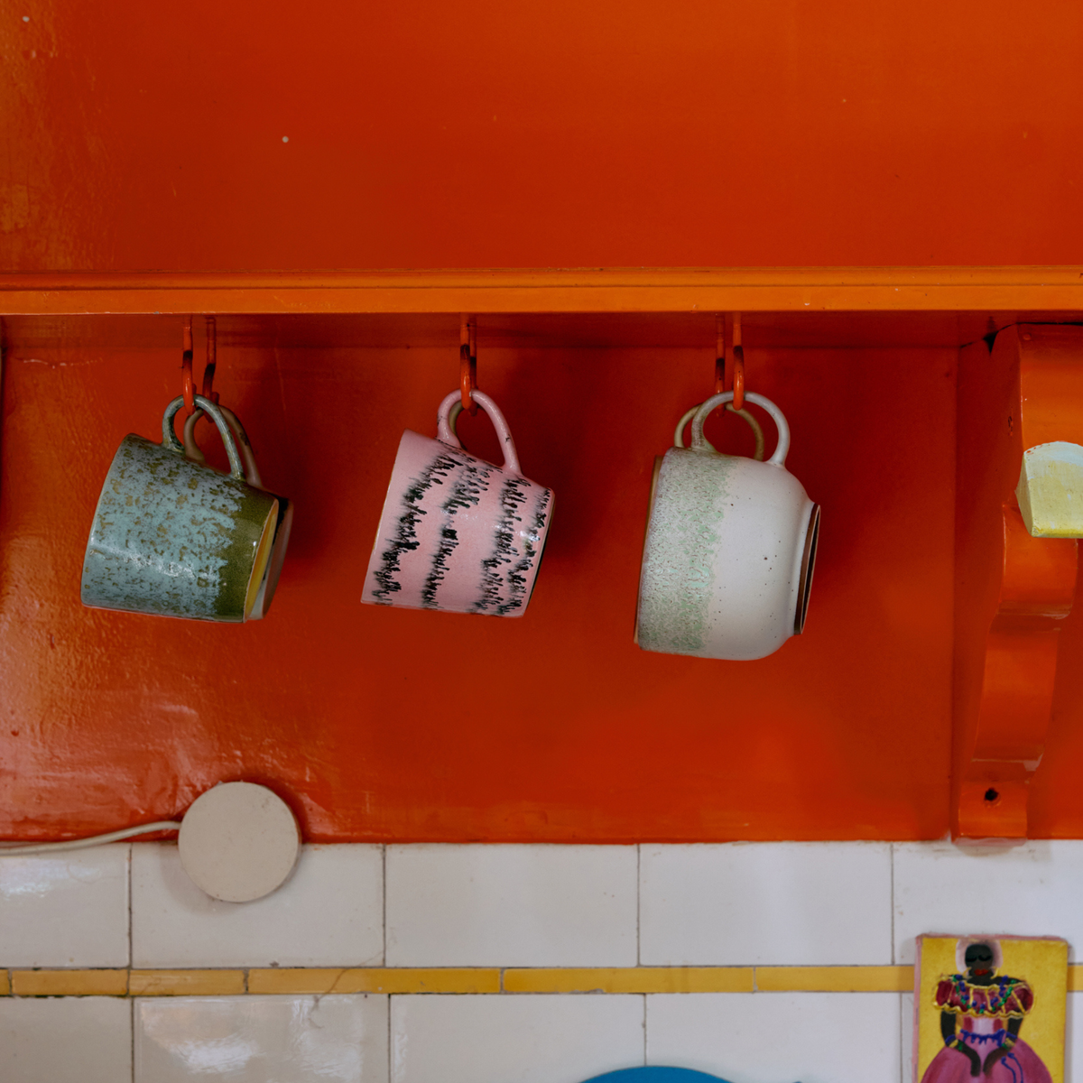 Drie HK Living 70s Ceramics Cappuccinokop Bubble mokken in groen, roze met zwarte lijnen en wit met groen hangen ondersteboven aan haken op een oranje keukenplank boven een betegelde muur.