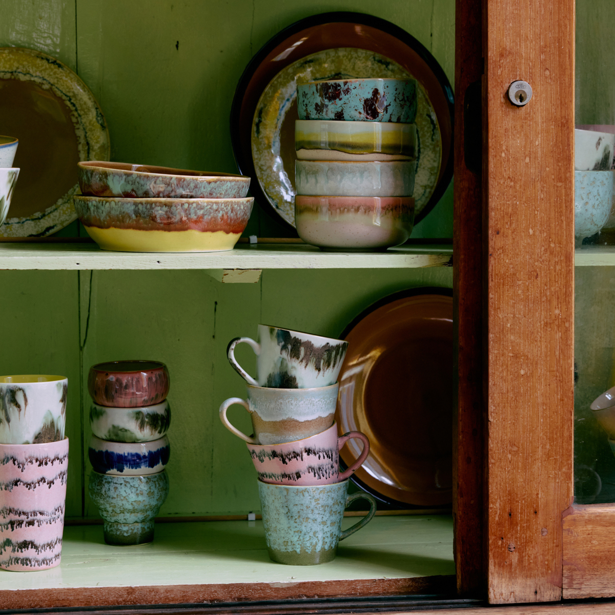 Een houten kast met glazen deuren toont kleurrijke HK Living 70s Ceramics Cappuccinokop Tide, kommen en borden met artistieke veelkleurige reactieve glazuren en patronen, netjes gestapeld op twee planken tegen een groene achtergrond.