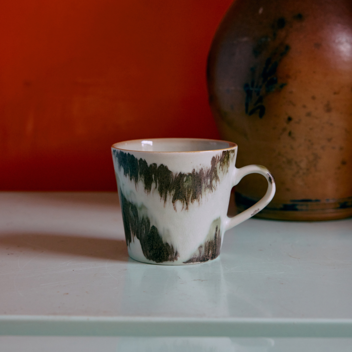 Een HK Living 70s Ceramics Cappuccinokop Tide met een bruin en wit abstract patroon rust op een lichte tafel. Een aardewerken pot is gedeeltelijk zichtbaar op de achtergrond tegen een oranje muur.