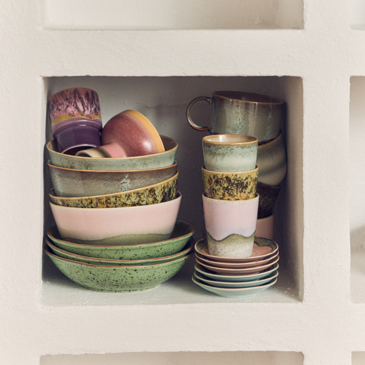 HK Living 70s Ceramics Koffiekop Petal en andere keramische schalen in gedempt groen, roze en aardetinten staan netjes gestapeld op een plank die is ingebouwd in een muur met witte textuur.