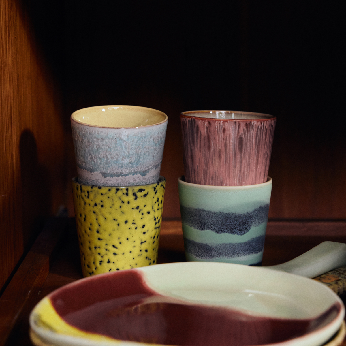 Vier HK Living 70s Ceramics Koffiemok Artistic mokken staan paarsgewijs op een houten plank naast een kom met een kastanjebruin en crèmekleurig patroon. Ze tonen de kleurrijke en gestructureerde stijl van HK Living's op de jaren 70 geïnspireerde keramiekcollectie.