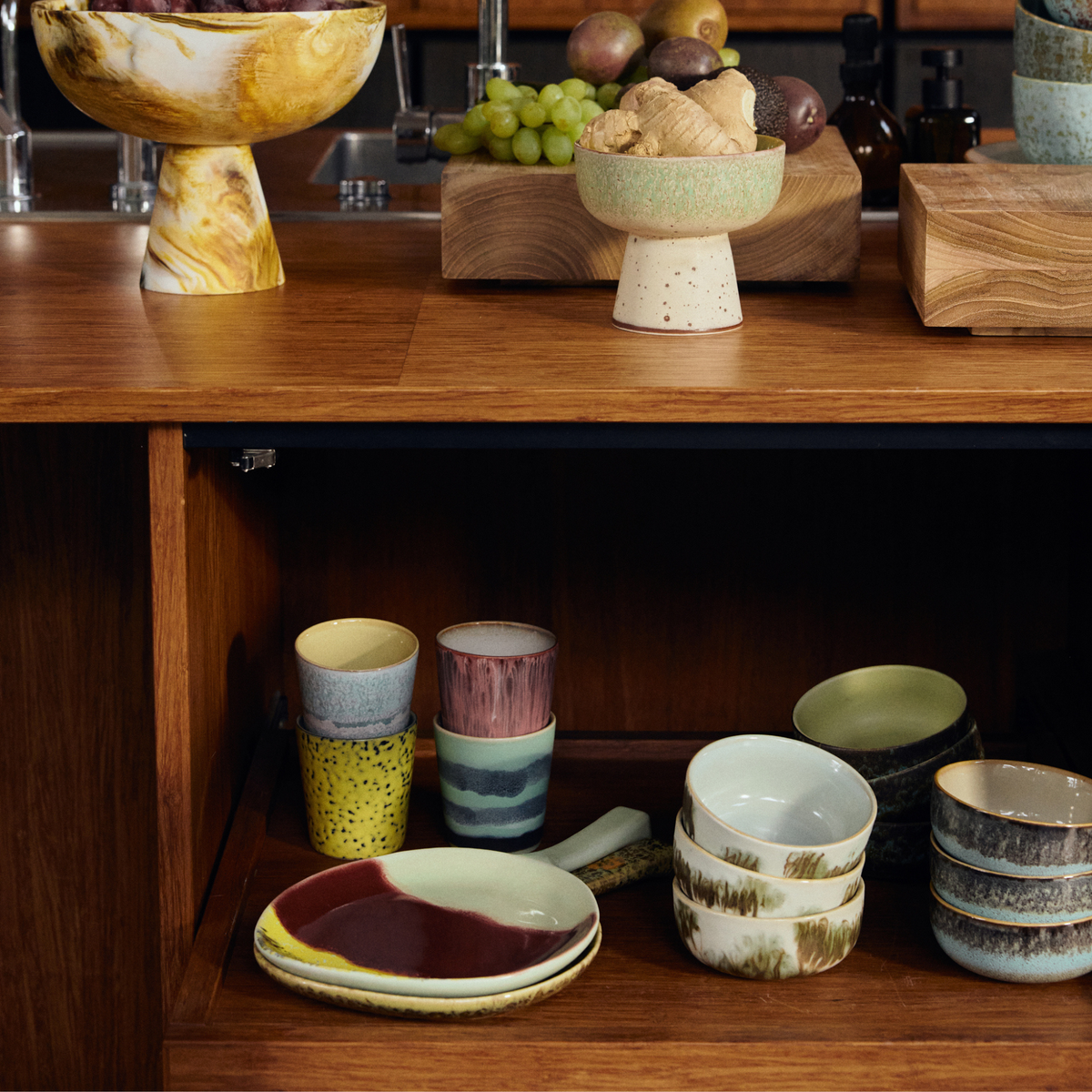 Een marmeren fruitschaal en een gemberschaal rusten op een houten toonbank boven planken met kleurrijke HK Living 70s Ceramics, waaronder de opvallende HK Living 70s Ceramics Koffiemok Artistic 4 st. mokken tussen levendige kopjes en kommen.