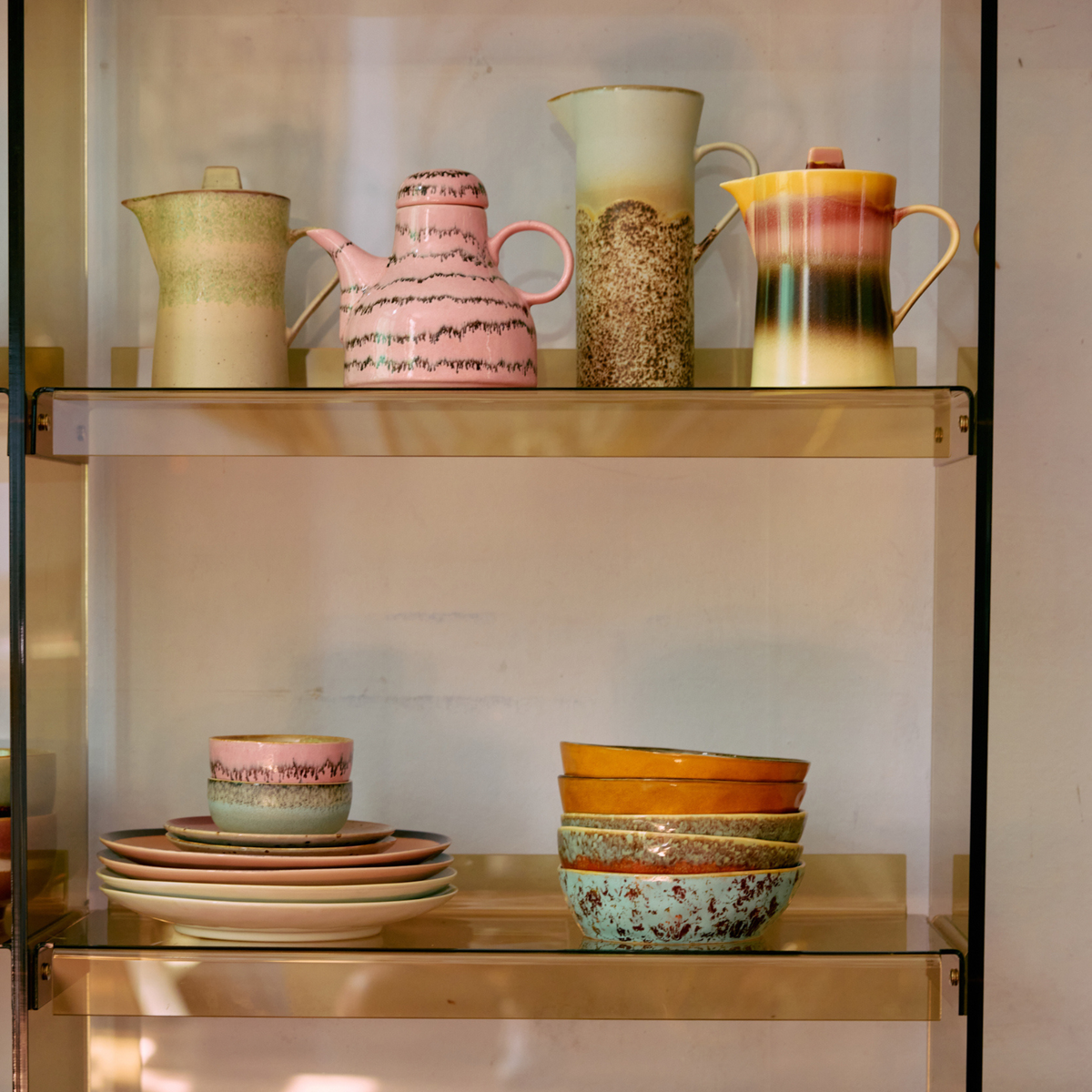 Op twee planken staat HK Living 70s Ceramics: drie theepotten en een hoge kan bovenop, terwijl eronder een stapel borden staat, een HK Living Pasta Kom Patina kom (set van 2), kop met schotel en meer in unieke pastelpatronen.