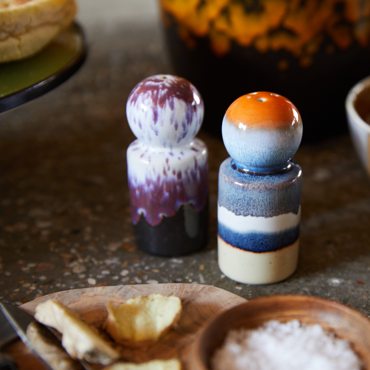 Twee HK Living 70s Keramiek Peper- en Zoutvaatje Stargaze op een tafel naast een houten schaal met zout en crackers. Retro tafelaccessoires en vaag aardewerk op de achtergrond maken dit charmante, kleurrijke tafereel compleet.