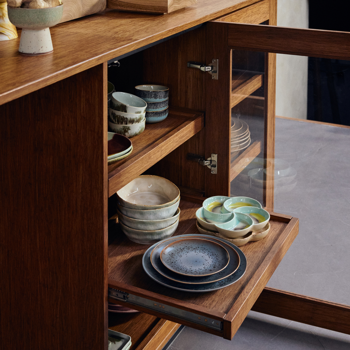 Een houten keukenkastje met glazen deuren gaat open en laat HK Living 70s Ceramics Pasta Kom Petal 2 st. kommen zien, netjes gerangschikt op planken, naast borden en mokken in aardetinten van HK Living.