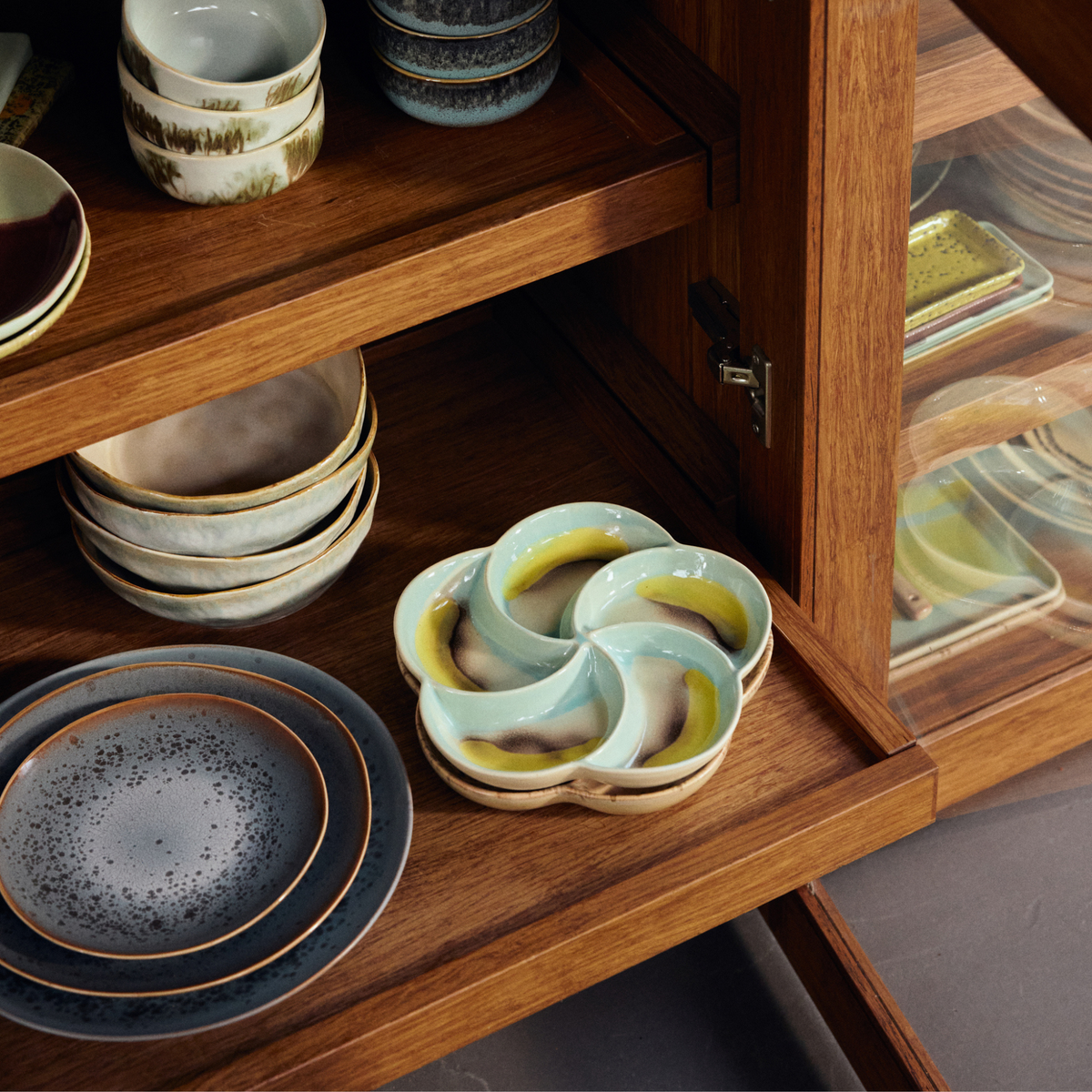 Een houten vitrinekast bevat HK Living 70s Ceramics Pasta Kom Petal 2 st., samen met gestapelde kommen, donker gespikkelde borden, een groenblauwe en gele bloemvormige schaal en geglazuurde rechthoekige borden, allemaal zichtbaar achter glazen deuren.