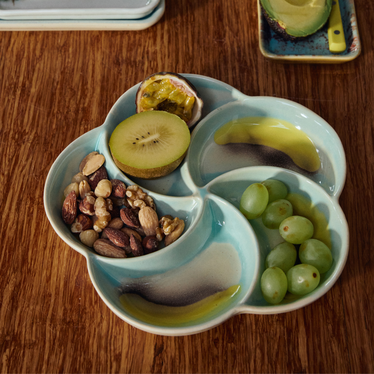De HK Living 70s Ceramics Snack Schaal Glint bevat gesneden kiwi, een halve passievrucht, groene druiven en verschillende noten op een houten tafel.