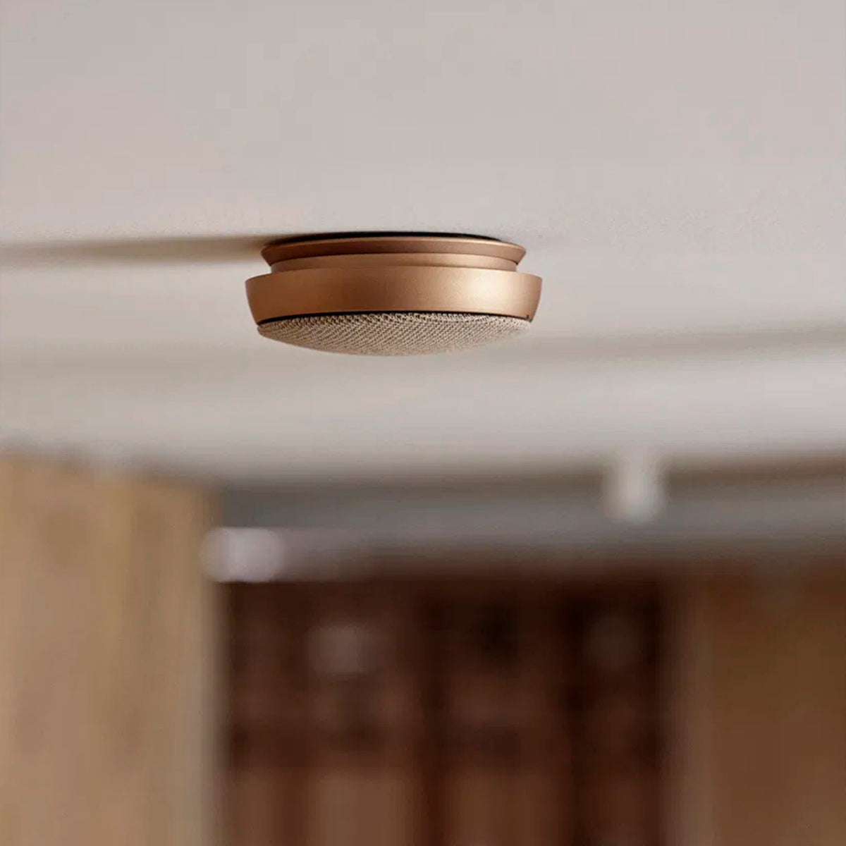 De Jacob Jensen Optische Rookmelder Koper D10 van Jacob Jensen is gemonteerd aan een wit plafond. Het koperkleurige ontwerp steekt elegant af tegen de vage achtergrond van een modern interieur.