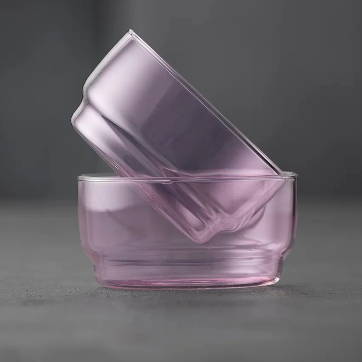 Twee Lyngby Glas Torino Kom Roze D12 kommen (set van 2) zijn schuin op elkaar gestapeld, waarbij de ene roze glazen kom diagonaal in de andere staat, tegen een effen grijze achtergrond.
