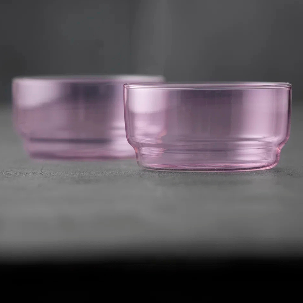 Twee transparante, lichtroze Lyngby Glas Torino Kom Roze D12 kommen (set van 2) rusten op een grijs oppervlak, de ene in focus terwijl de andere zachtjes naar de achtergrond verdwijnt. Merk: Lyngby Glas.