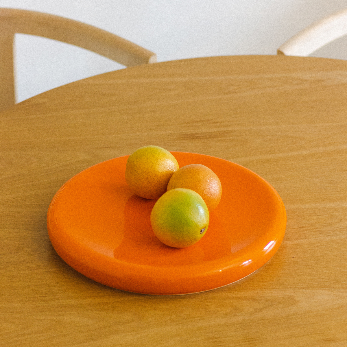 Drie sinaasappels liggen op een „Big O Serveerschaal Orange“ van Matias Moellenbach, die op een lichte houten tafel staat, met op de achtergrond een houten stoel met een gebogen rugleuning.