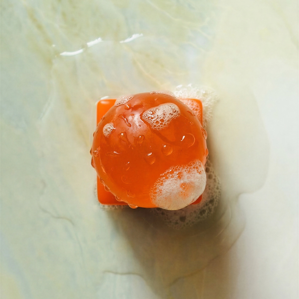 Een oranje bol, die doet denken aan een Matias Moellenbach OBA Zeep Bar Papaya, is bedekt met zeepbellen en staat op een vierkante oranje voet, net als een navulzeep OBA Zeep Houder. Rondom liggen water en zeepsop op een bleek, glad oppervlak.