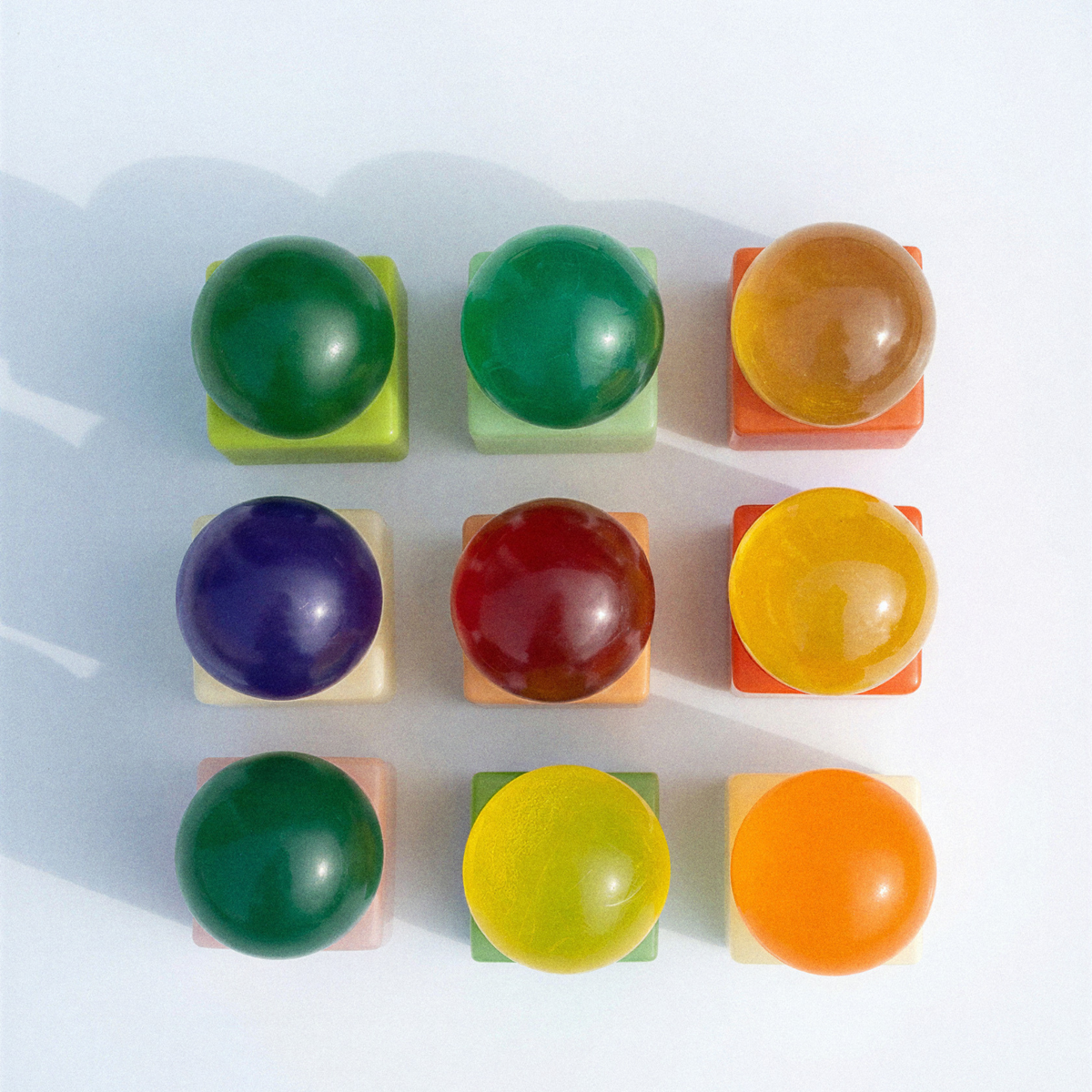 Negen levendige, doorschijnende bollen, elk geplaatst op een vierkante pastelkleurige voet en gerangschikt in een raster van 3 bij 3, weerspiegelen de speelse esthetiek van de Matias Moellenbach OBA Zeep Bar Aloe Vera. Elke bol heeft een unieke tint – waaronder groen, geel, oranje, rood en paars – die aansluit bij de levendige kleuren van de vaste zeep van Matias Moellenbach en zachte schaduwen werpt op een witte achtergrond.