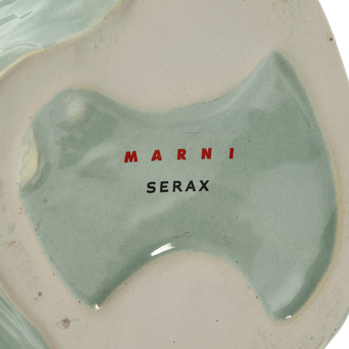 De basis van de Serax Marni After Midnight Uil Vaas Lichtblauw XS vaas van keramiek heeft een glanzend lichtblauw glazuur en op het ongeglazuurde gedeelte staat "MARNI" in rode en "SERAX" in zwarte letters.