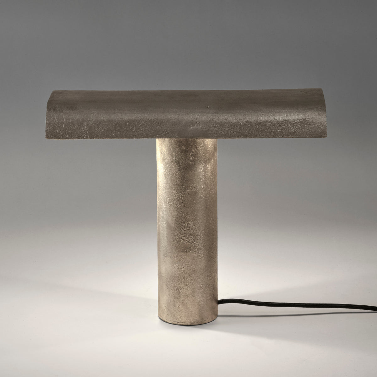 De Serax Fck Tafellamp Staal Grijs van Serax heeft een modern, minimalistisch ontwerp met een cilindrische voet, platte rechthoekige bovenkant, afwerking met metallic textuur en zichtbaar zwart netsnoer op een wit oppervlak.